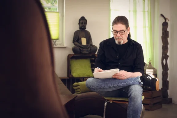 Ein Mann sitzt auf einem Stuhl, liest Notizen, im Hintergrund eine Buddha-Statue.