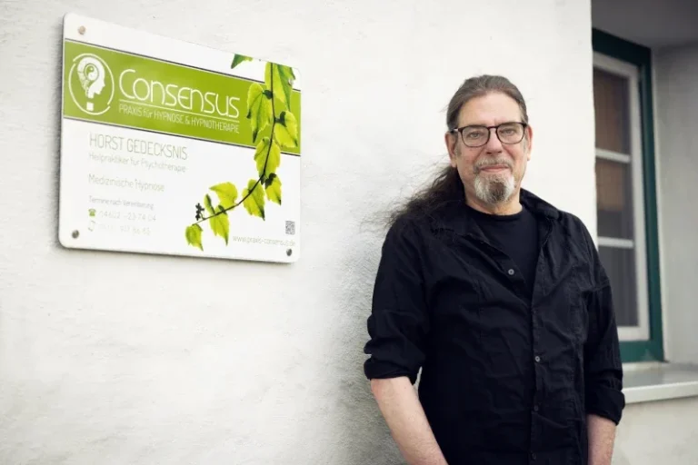 Hypnose Praxis Consensus Großsolt Mann mit Brille und langen Haaren steht neben einem Schild mit Firmenlogo und Pflanzenmotiven.